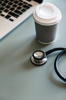 A picture of a grey take-away coffee cup next to a laptop and a stethoscope on a grey table